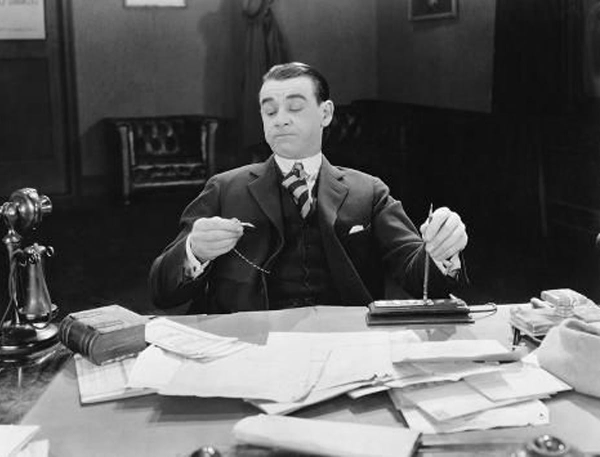 A man in a suit sits at a cluttered desk, leaning back with a smug expression, holding a pocket watch chain in both hands. Papers, a telephone, and books are scattered across the desk.