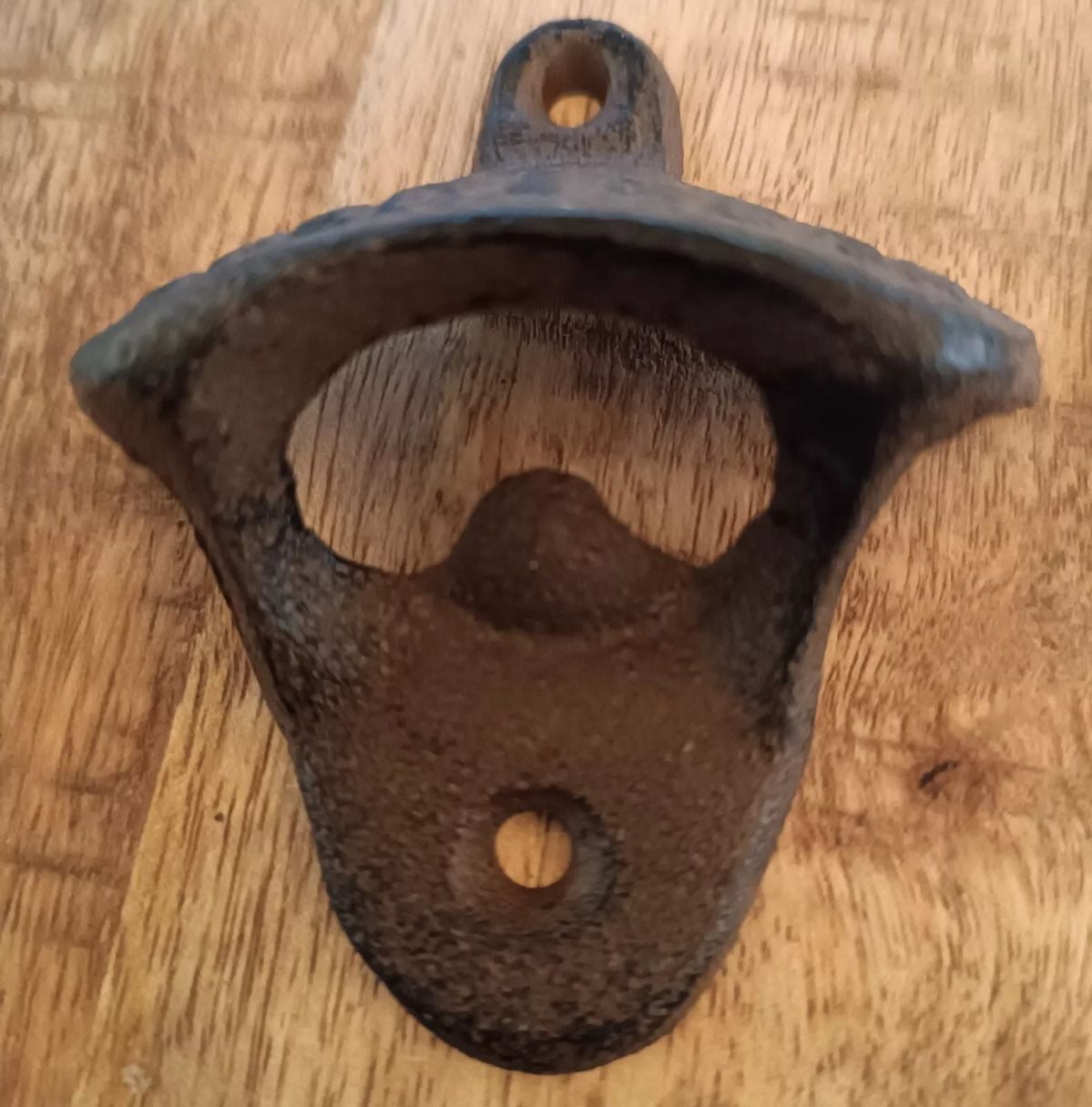 A close-up of a metal wall-mounted bottle opener with two screw holes, attached to a wooden surface. The opener has a curved lip for prying open bottle caps.