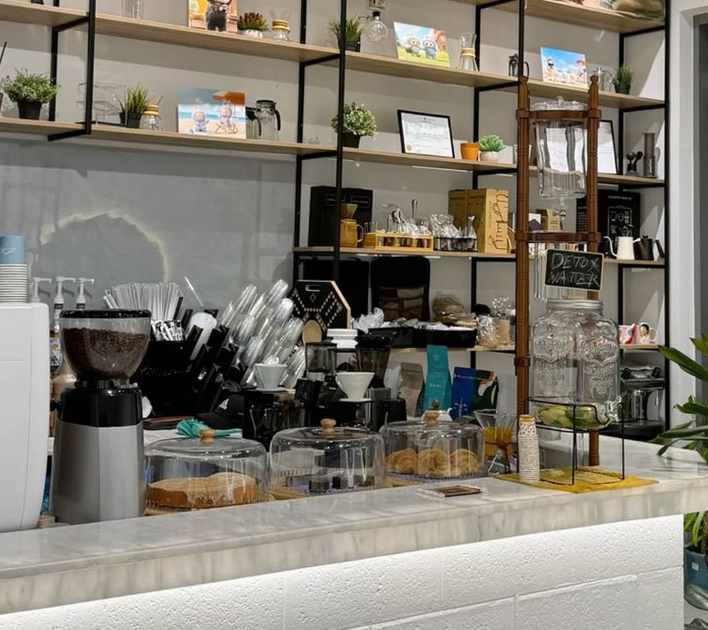 A modern café counter with a coffee grinder, glass-covered pastries, jars of detox water, and shelves displaying mugs, plants, and décor against a light wall. The space appears clean and organized.