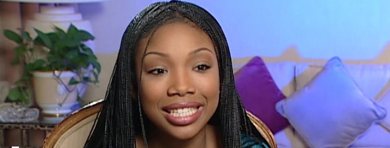 A young woman with long braided hair smiles while sitting indoors. Behind her are a white lamp, a green plant, and several pastel-colored pillows on a couch. The background is softly lit with blue and purple tones.