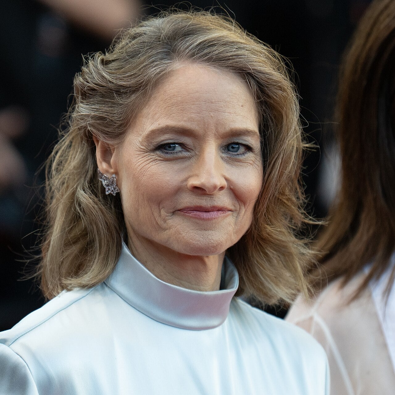 A woman with shoulder-length light brown hair, styled in soft waves, wears a high-collared, pale satin top and star-shaped earrings, smiling slightly at a formal event.