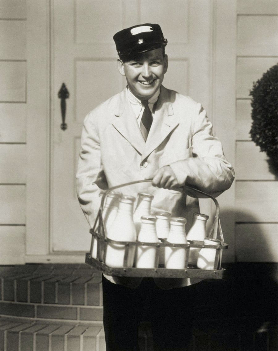 A smiling milkman dressed in a uniform and cap stands on a doorstep, holding a basket filled with glass milk bottles, in front of a house with brick steps and a wooden door.