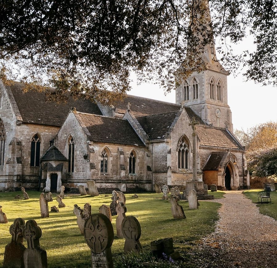 A historic stone church with a tall steeple stands amid a graveyard filled with old headstones and shaded by large trees, bathed in soft afternoon sunlight.