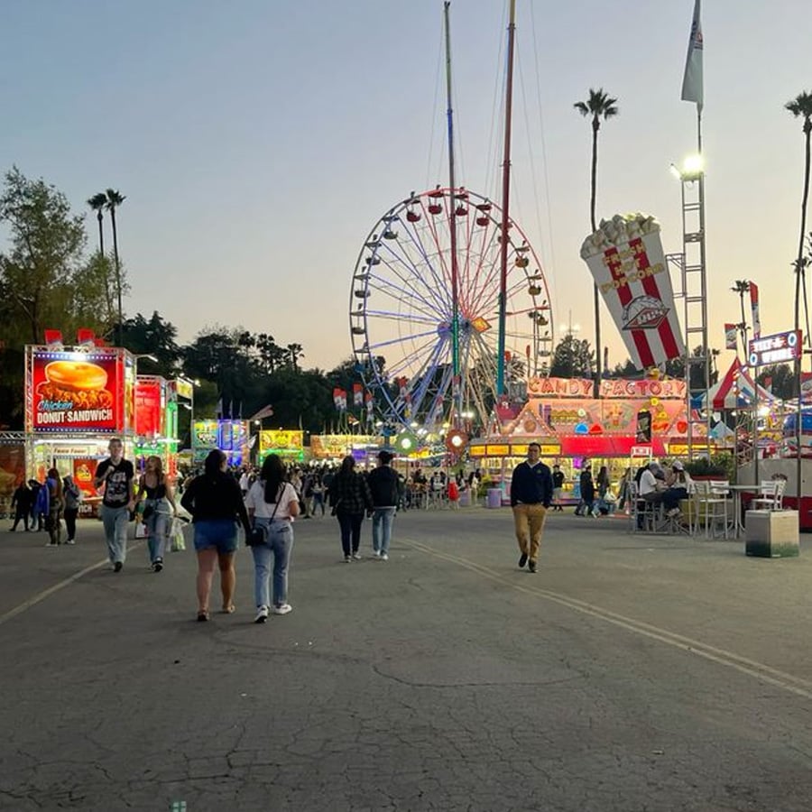 People walk through a lively fairground at sunset, with food stalls, bright signs, and a large Ferris wheel in the background. Tall palm trees line the area and the sky is clear.