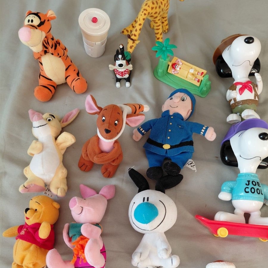 A collection of small stuffed toys and figurines, including characters like Tigger, Winnie the Pooh, Piglet, Snoopy, and a blue-uniformed doll, arranged on a light-colored surface.