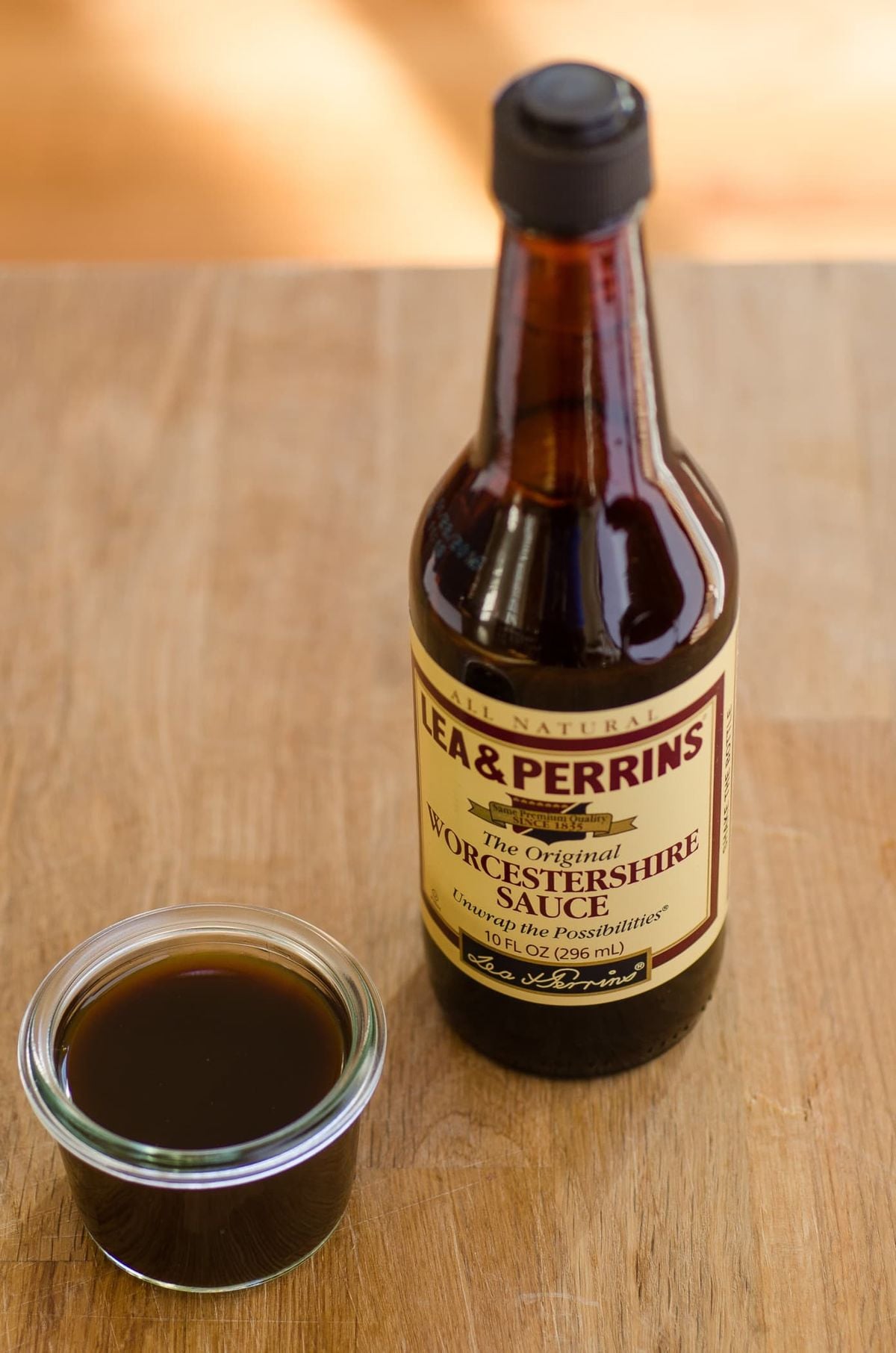 A bottle of Lea & Perrins Worcestershire Sauce stands on a wooden surface next to a small glass dish filled with dark brown sauce.