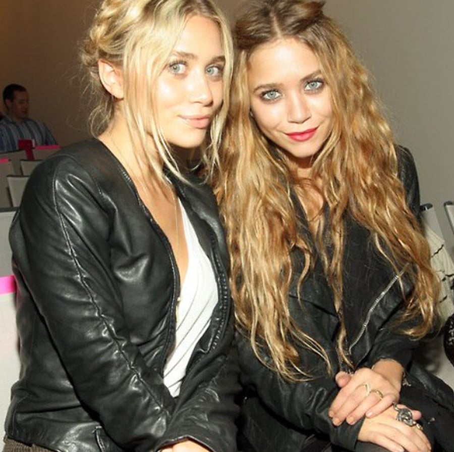 Two women with long hair and light eyes sit close together, both wearing black leather jackets and light-colored tops, smiling at the camera in a bright indoor setting.