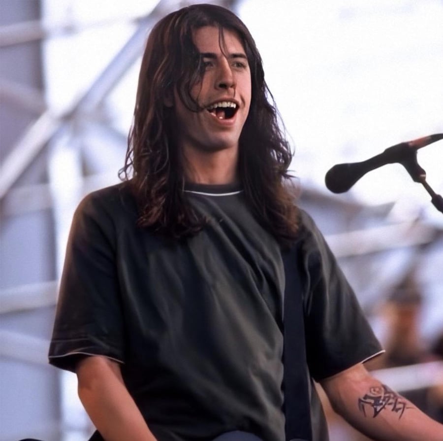 A musician with long dark hair smiles while performing on stage, wearing a dark t-shirt. A microphone is positioned nearby, and a tattoo is visible on the person's forearm. The background shows a blurred stage structure.