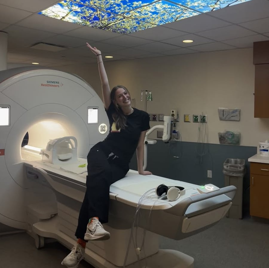 A smiling woman in black clothes sits on an MRI machine, raising one arm toward a ceiling panel with a nature scene. The room has medical equipment, cabinets, and soft lighting.