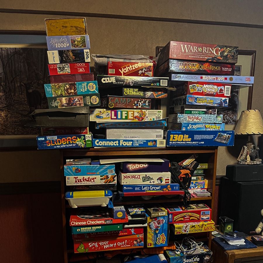 A shelf stacked with various board games, including The Game of Life, Risk, Connect Four, Taboo, Twister, Chinese Checkers, and more, in a cozy room with brown walls and a framed photo on the wall.