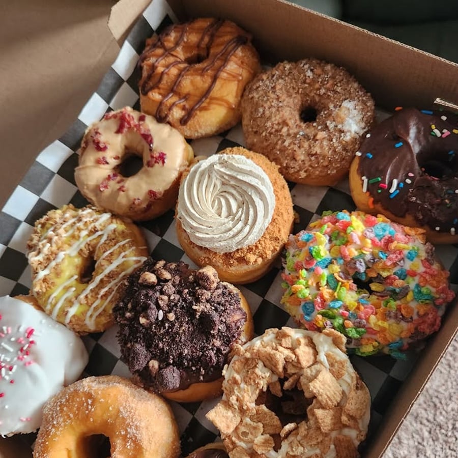 A box of twelve assorted donuts with various colorful toppings such as chocolate, sprinkles, crushed cookies, cereal, and icing, arranged on a black-and-white checkered paper.