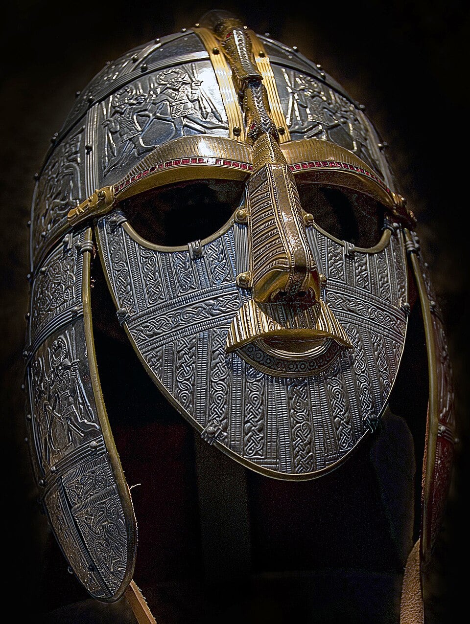 An ornate, silver and gold Anglo-Saxon helmet with intricate engravings and patterns, featuring a prominent nose guard and red accents, shown against a dark background.