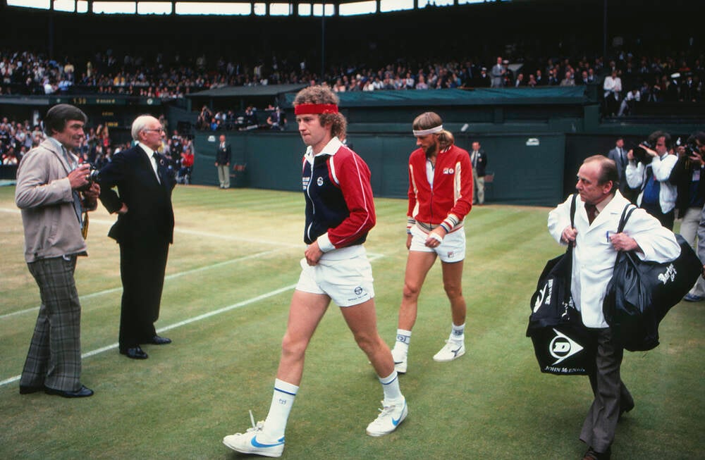 Two male tennis players walk off a grass court at Wimbledon, surrounded by officials and photographers, while a crowd watches from the stands. The players wear vintage tennis outfits and one carries a tennis bag.