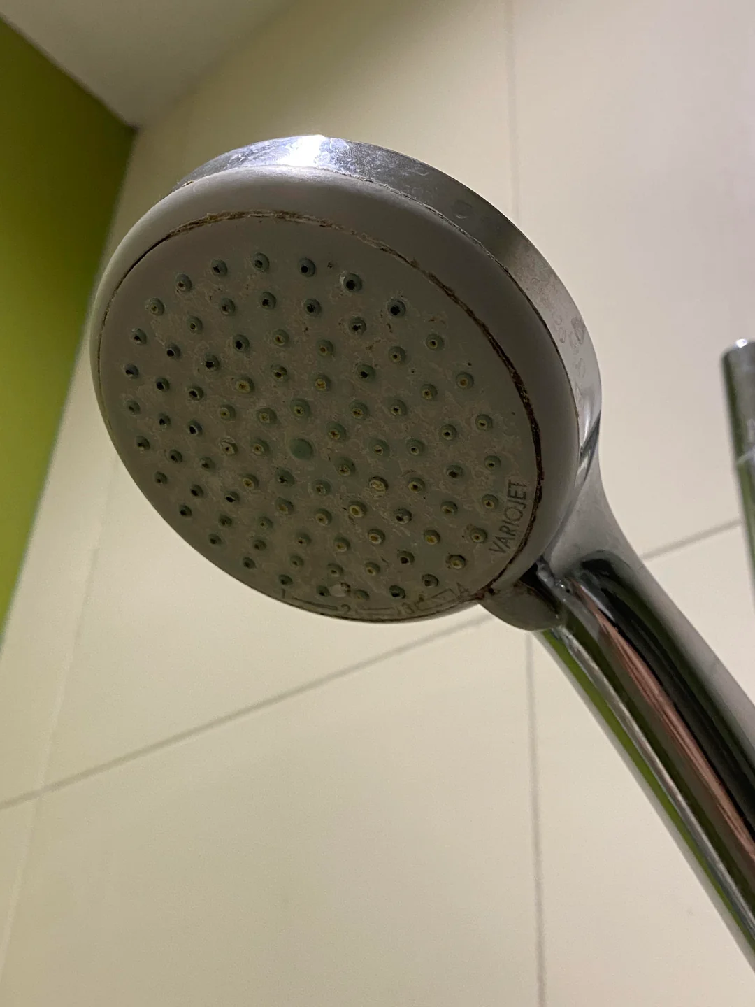 Close-up view of a metallic handheld showerhead mounted on a tiled bathroom wall, with some minor rust or mineral buildup visible around the edges.