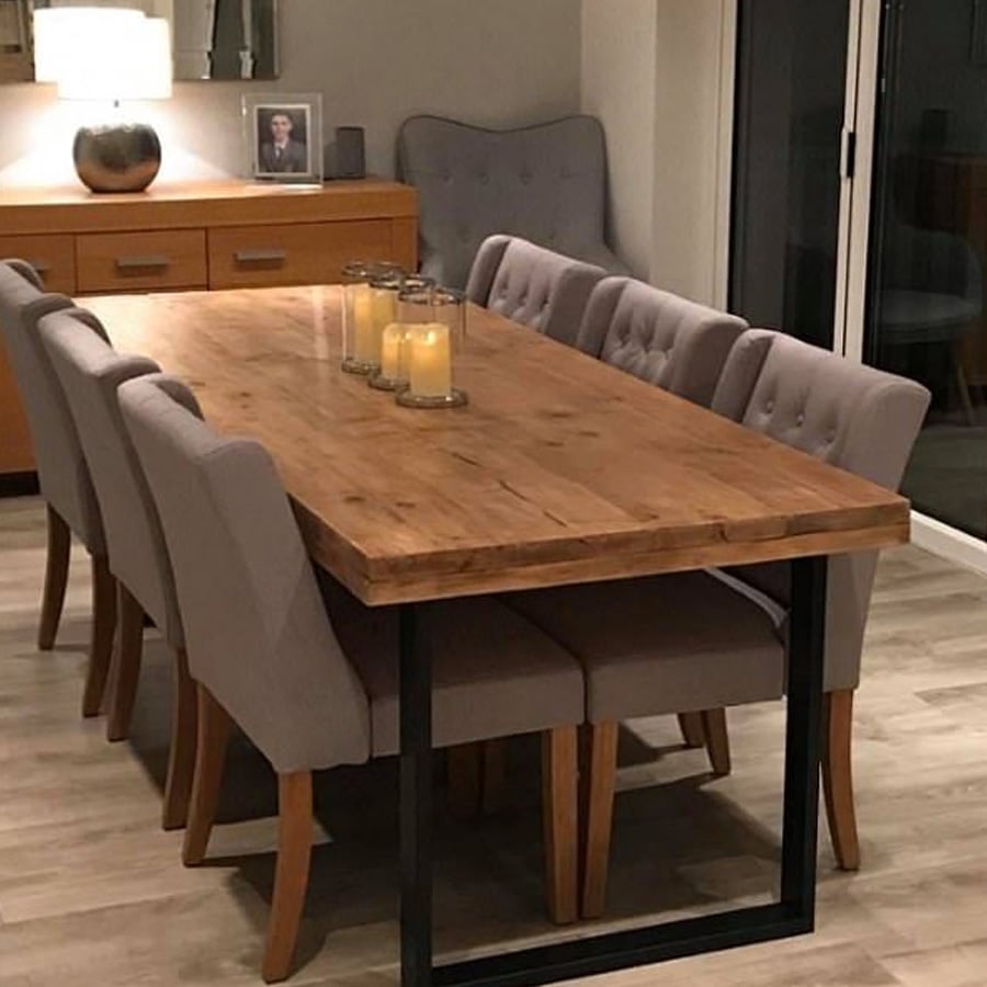 A dining room with a wooden table, eight gray upholstered chairs, and four candles in glass holders. In the background are a sideboard, gray armchair, lamp, decorative vase, and framed photo.