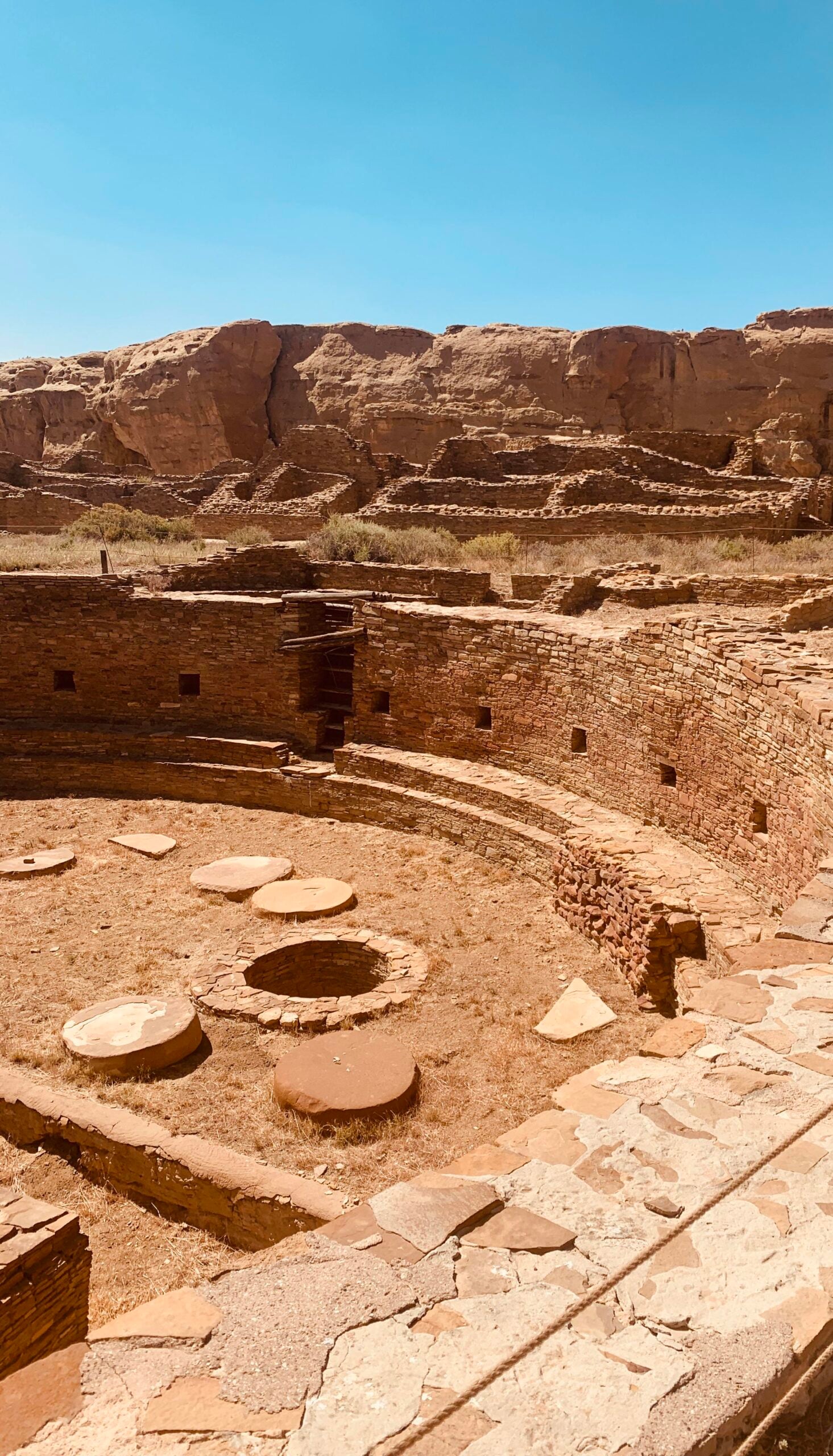 Ancient circular stone ruins with stepped walls, stone slabs, and a central pit, set against a backdrop of rocky cliffs under a clear blue sky.