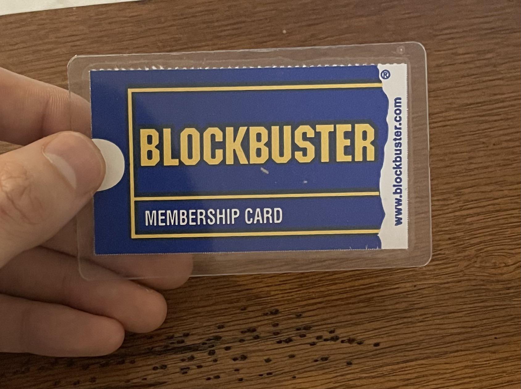 A hand holds a plastic-covered Blockbuster membership card, featuring the blue and yellow Blockbuster logo and the website address, against a wooden background.