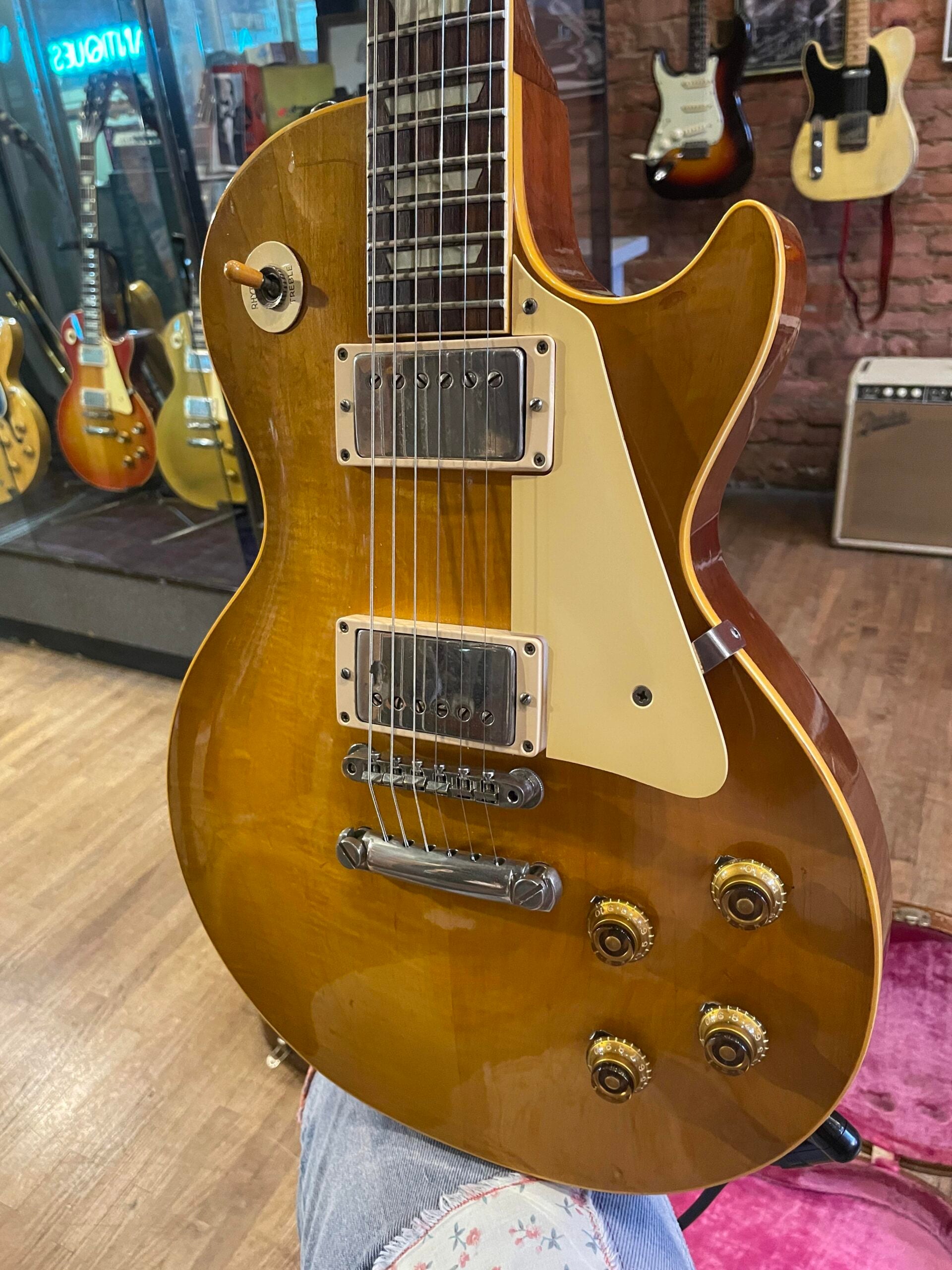 A close-up of a sunburst electric guitar with a glossy finish, four control knobs, and two humbucker pickups, displayed in a music store with other guitars and an amplifier in the background.