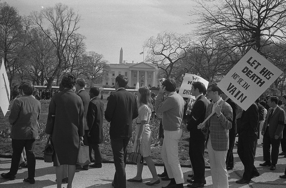 Black and white photo of a group of people marching near the White House, holding signs. One sign reads “LET HIS DEATH NOT BE IN VAIN.” The Washington Monument is visible in the background.