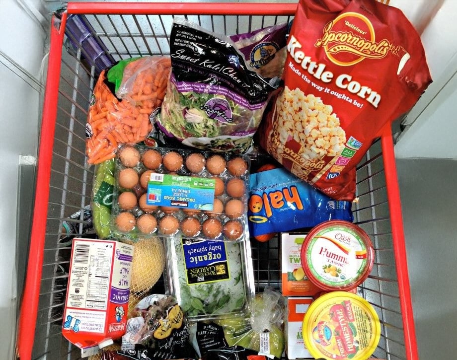 A shopping cart filled with groceries including eggs, kettle corn, baby carrots, salad mix, organic greens, hummus, Halo mandarins, and packaged food items, viewed from above.