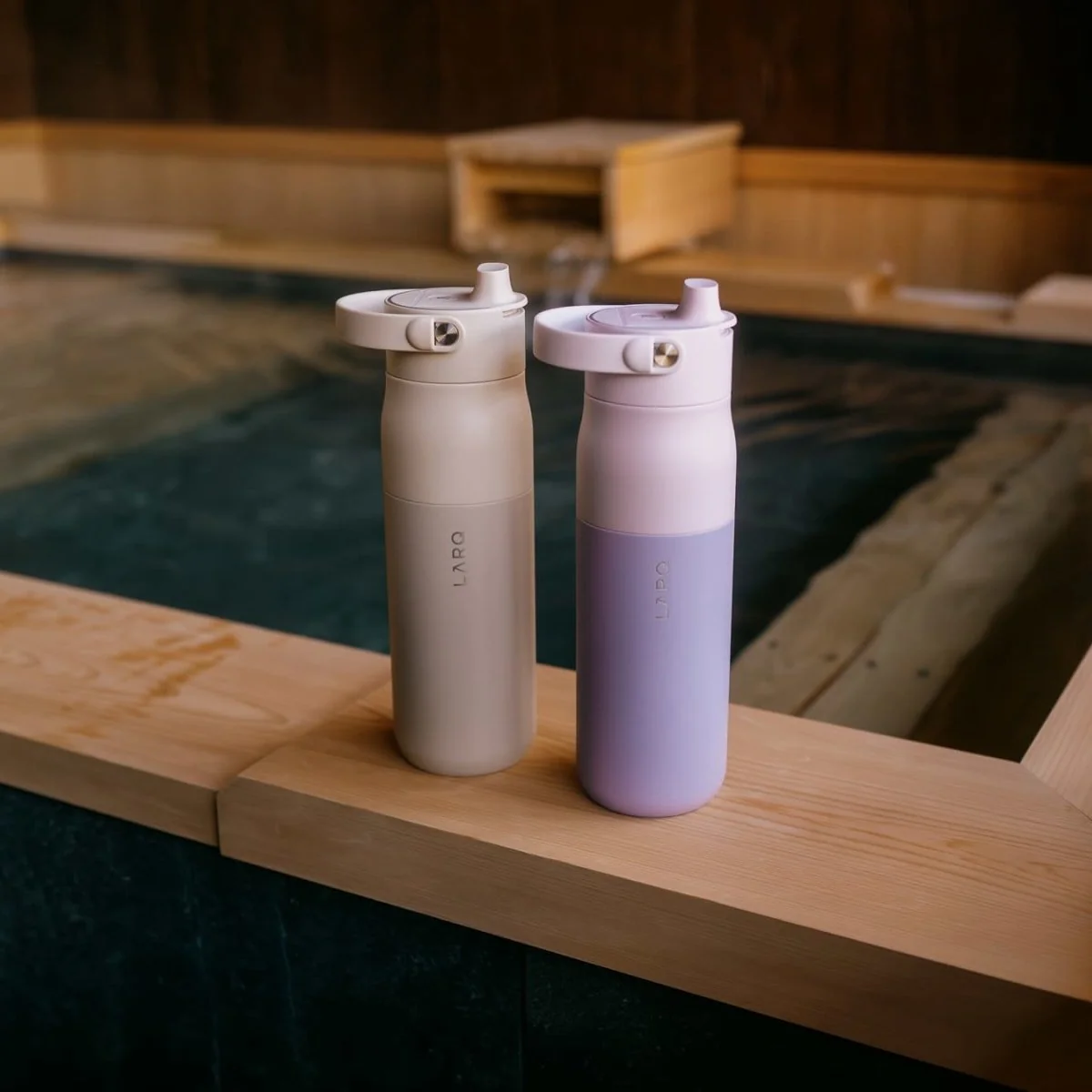 Two reusable LARQ water bottles, one beige and one lavender, stand side by side on the wooden edge of a bath or pool, with water and wooden structures visible in the background.