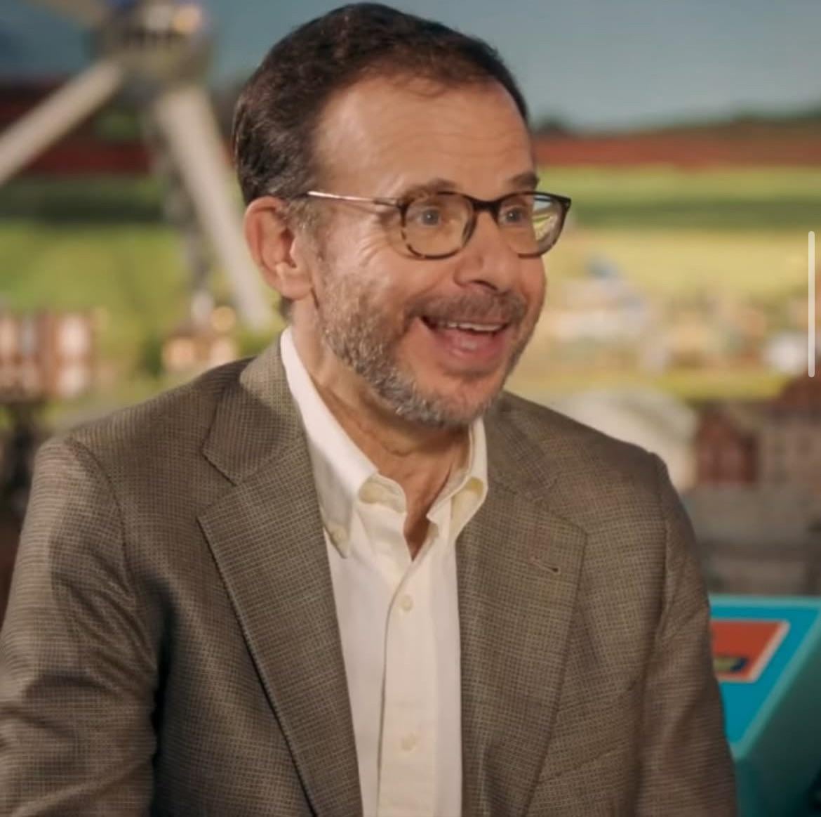 A man with short brown hair, glasses, and a beard smiles while wearing a brown checked blazer and a white shirt, with a colorful, blurred background behind him.