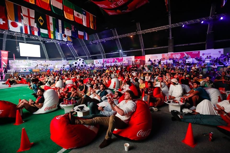 A large crowd sits on red and white bean bags in an indoor arena, watching a sporting event on a big screen. International flags hang from the ceiling; Coca-Cola branding is visible throughout the venue.