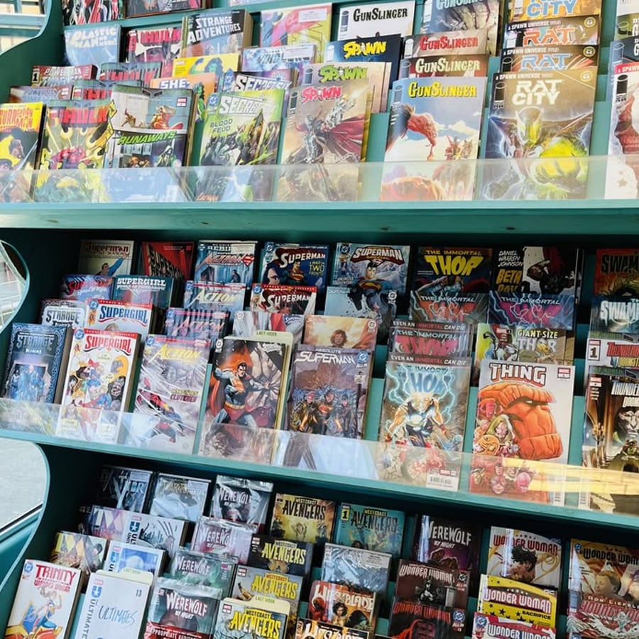 A display shelf filled with various colorful comic books, including titles like Superman, Spawn, The Thing, Thor, and The Avengers, organized in rows at a comic book store.