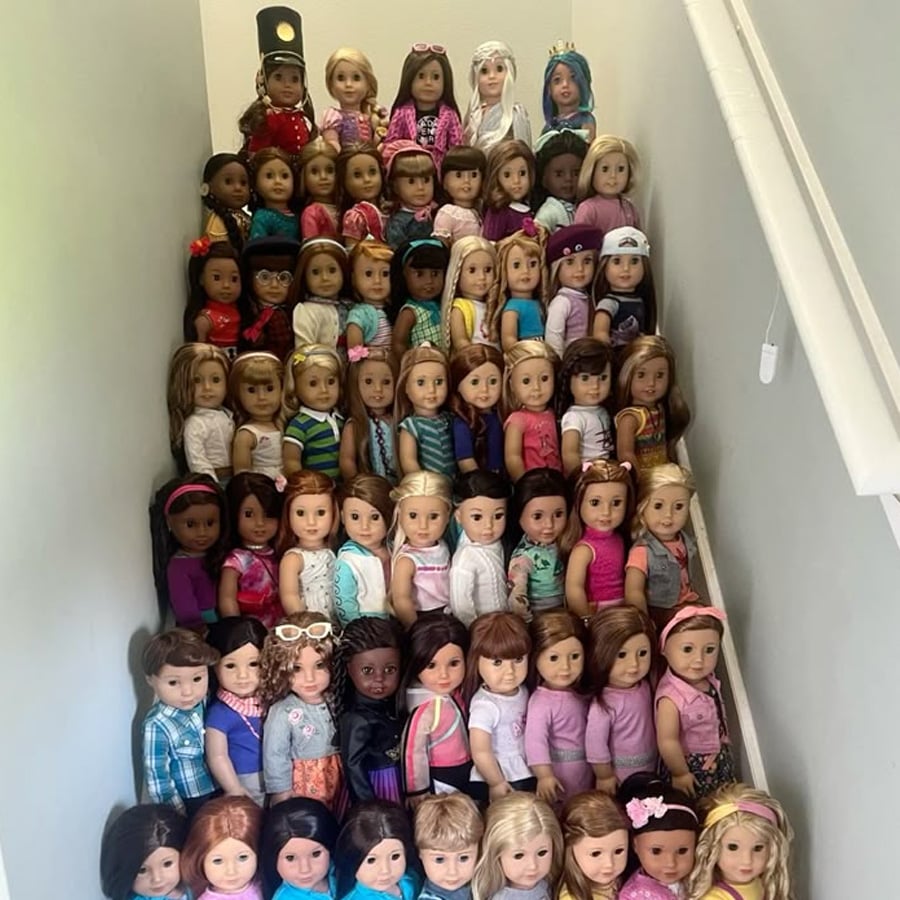 Over fifty dolls, dressed in a variety of colorful outfits, are neatly arranged sitting on the steps of a staircase, filling the entire space from top to bottom. The dolls have different hairstyles and skin tones.