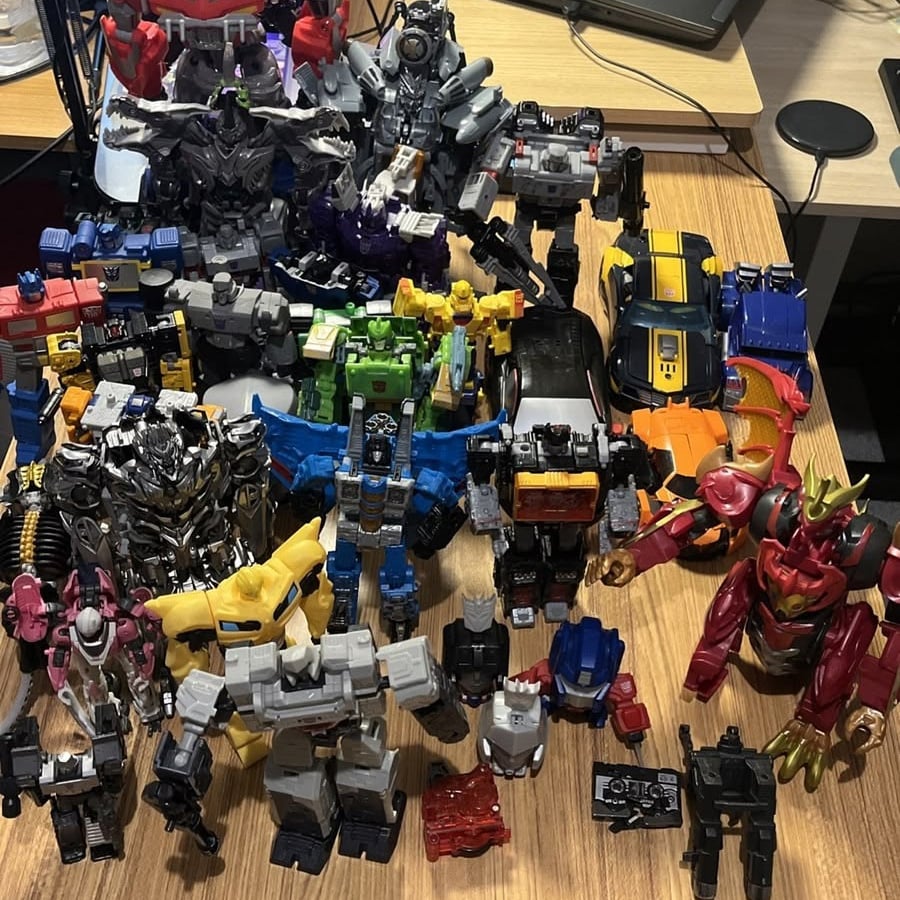 A collection of various Transformers action figures, both robot and vehicle forms, are arranged closely together on a wooden desk with electronics and other items in the background.