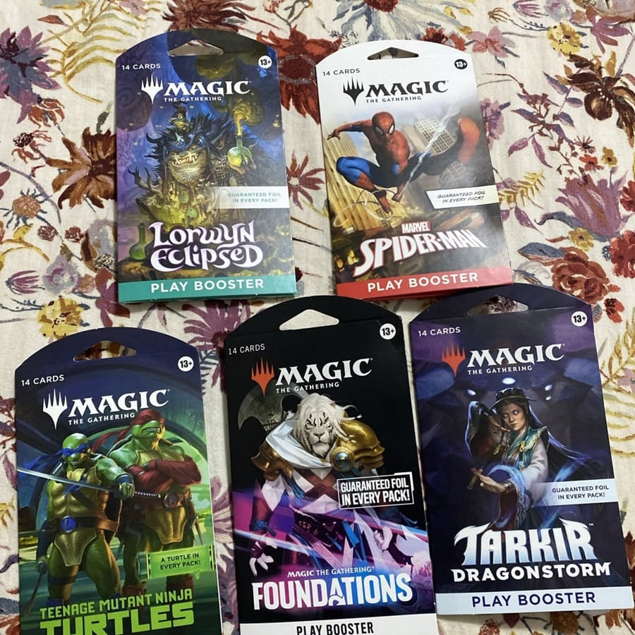 Five Magic: The Gathering play booster packs, including Lorwyn Eclipsed, Marvel Spider-Man, Teenage Mutant Ninja Turtles, Foundations, and Tarkir Dragonstorm, arranged on a floral-patterned fabric.