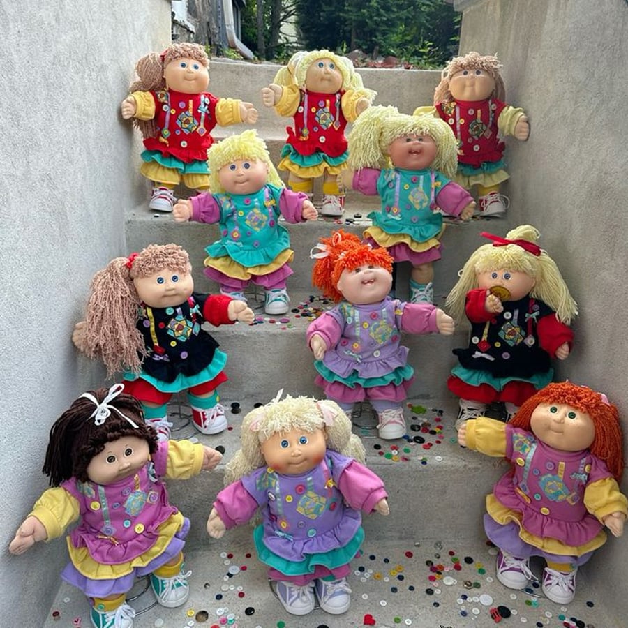 Several Cabbage Patch Kid dolls with yarn hair in colorful dresses stand on outdoor concrete steps covered with confetti. The dolls have round faces, wide eyes, and various hair colors and styles.
