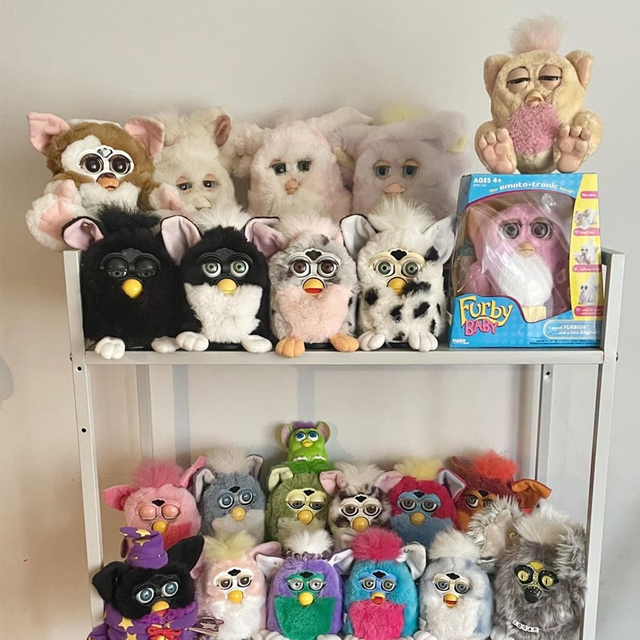 A white shelf displays a collection of vintage Furby and Gizmo plush toys, including various colors and styles. One Furby is in its original box. The toys are neatly arranged in two rows.