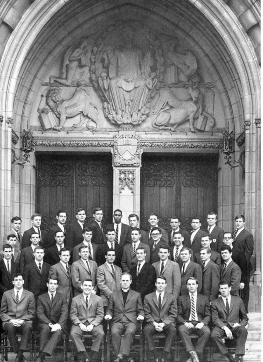 A black-and-white photo of a large group of men in suits posing for a formal portrait on the steps of a building with ornate stone carvings and arched doorways in the background.