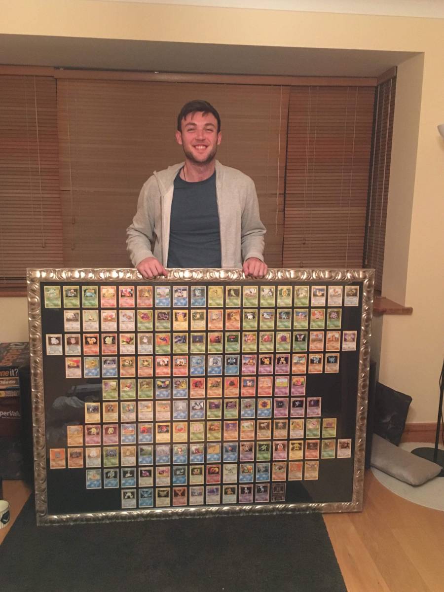 A smiling man stands indoors holding a large, framed display filled with numerous Pokémon trading cards arranged in a grid. The display appears to be a complete collection of the original 151 Pokémon.