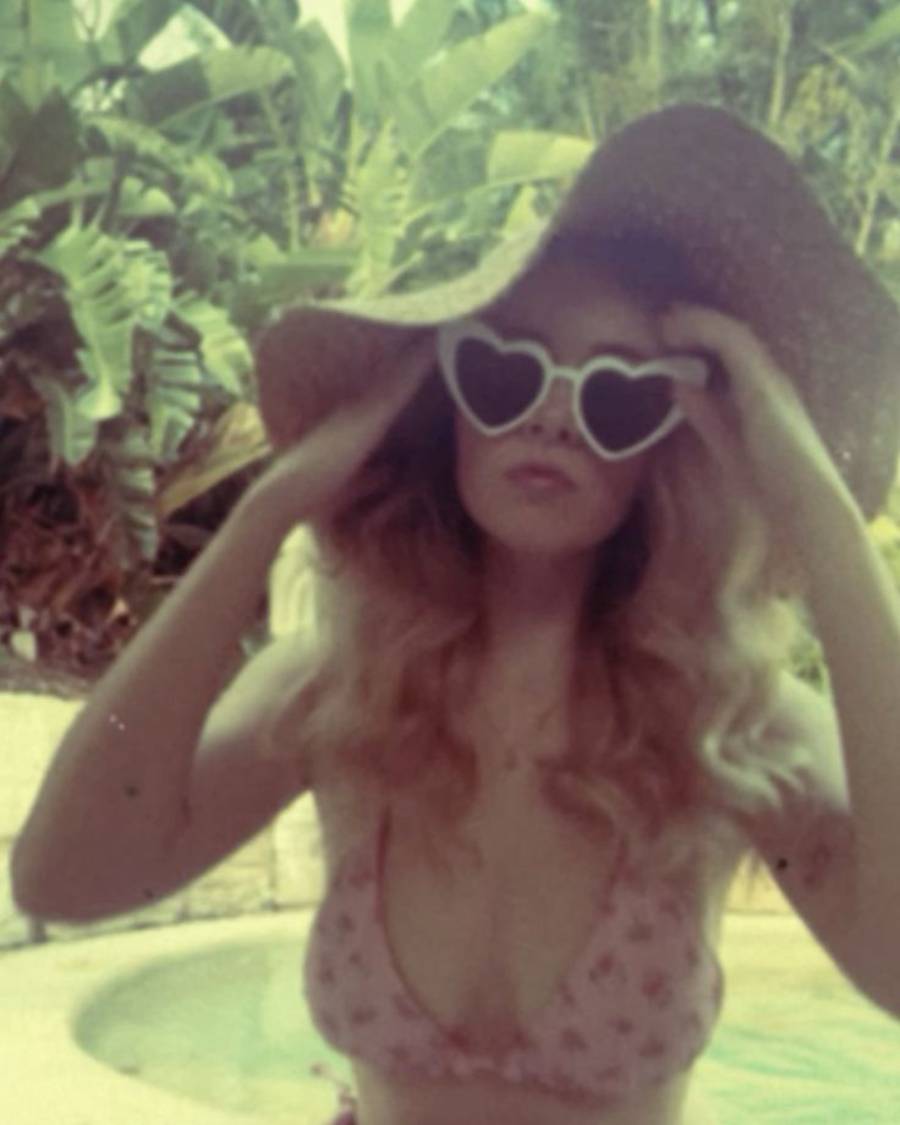 A woman in a bikini, heart-shaped sunglasses, and a wide-brimmed hat stands by a pool surrounded by lush tropical plants.