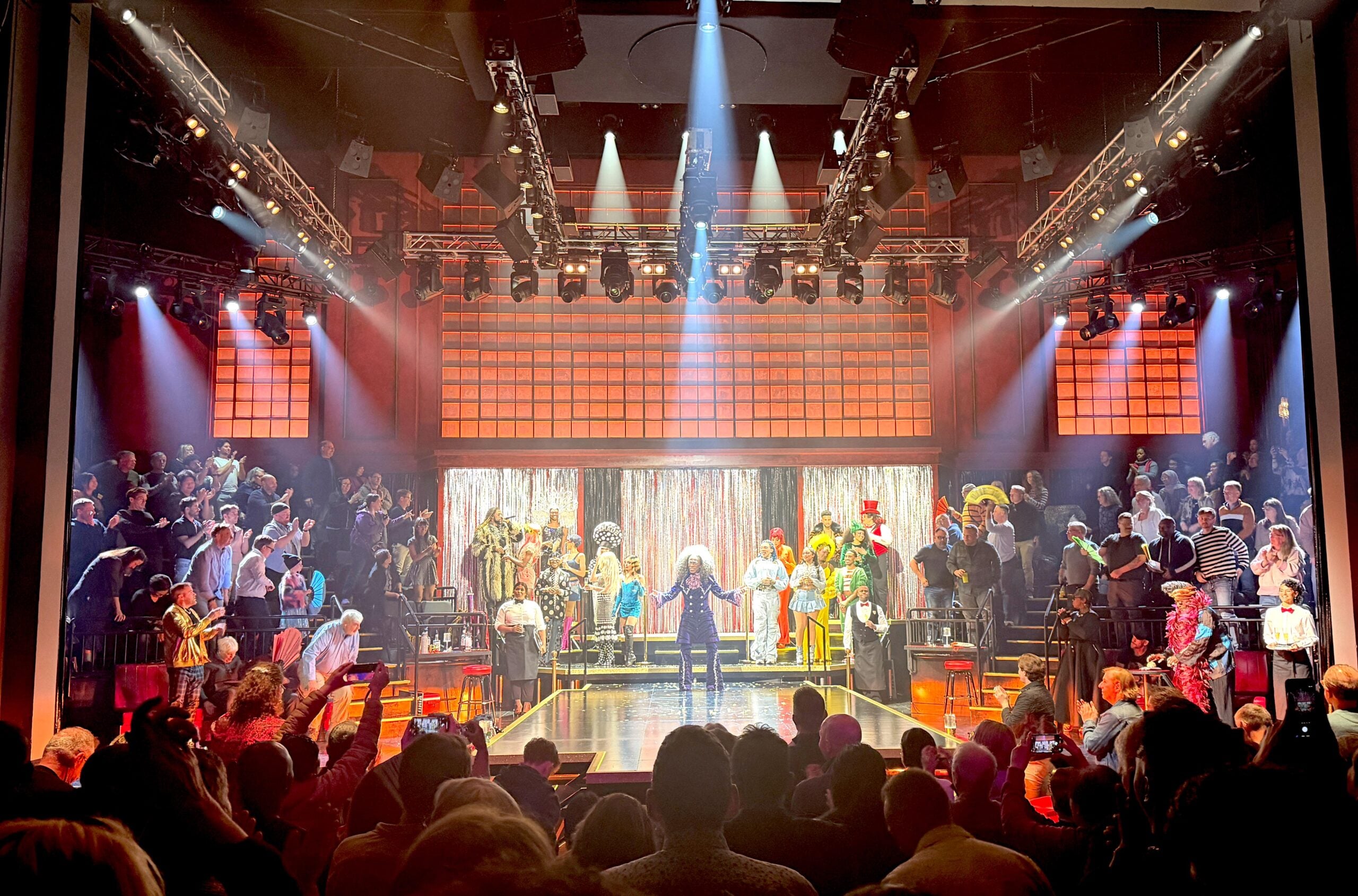 A vibrant stage performance with a singer in the spotlight at center stage, surrounded by performers in colorful costumes. The audience sits on both sides, watching the lively theater show under dramatic lighting.