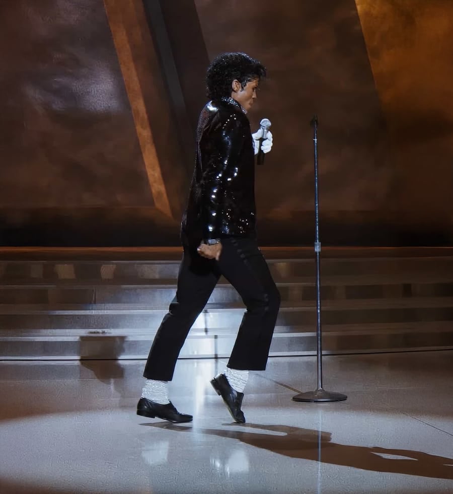 A person in a black sequin jacket, black pants, white socks, and black shoes performs a dance move on stage near a microphone stand, with a spotlight and brown background behind them.