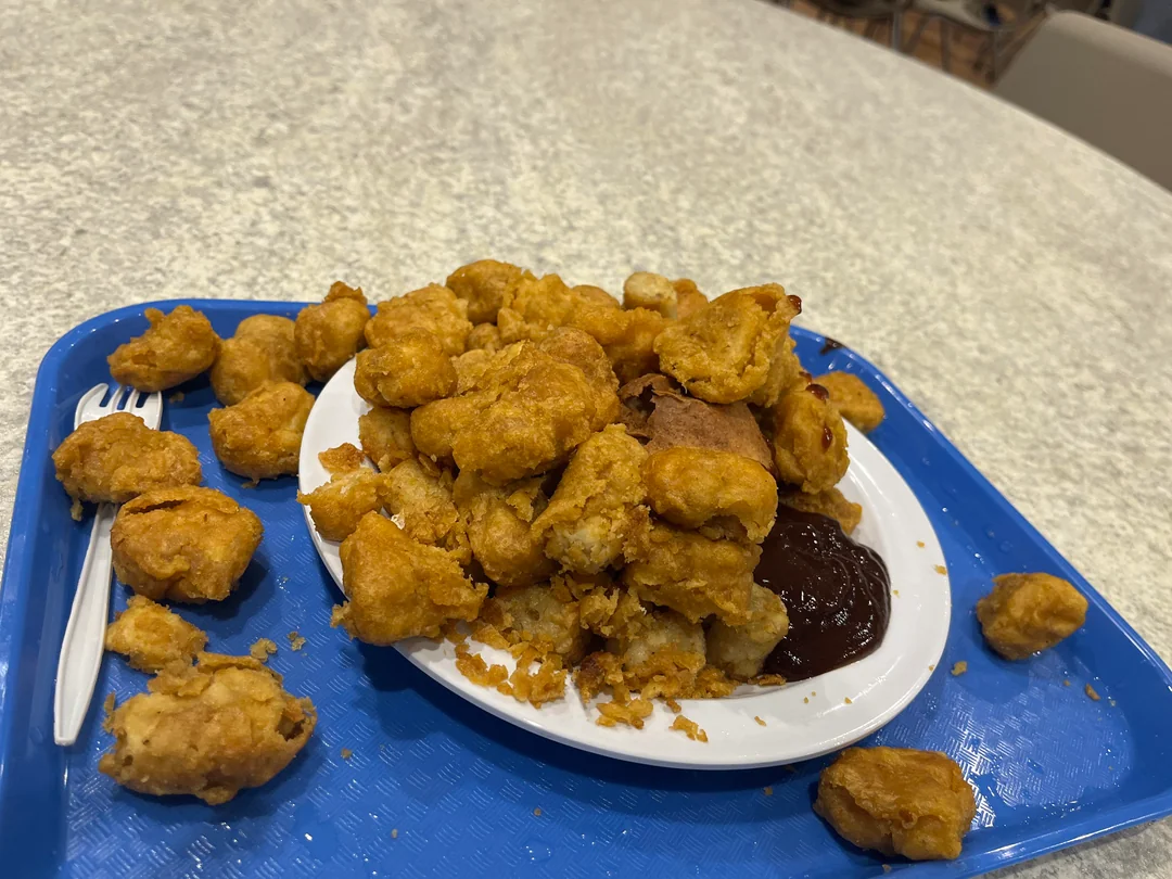 A plate piled with golden-brown, fried nuggets and a serving of barbecue sauce sits on a blue tray. Some nuggets have fallen onto the tray, and a plastic fork is placed on the left side.