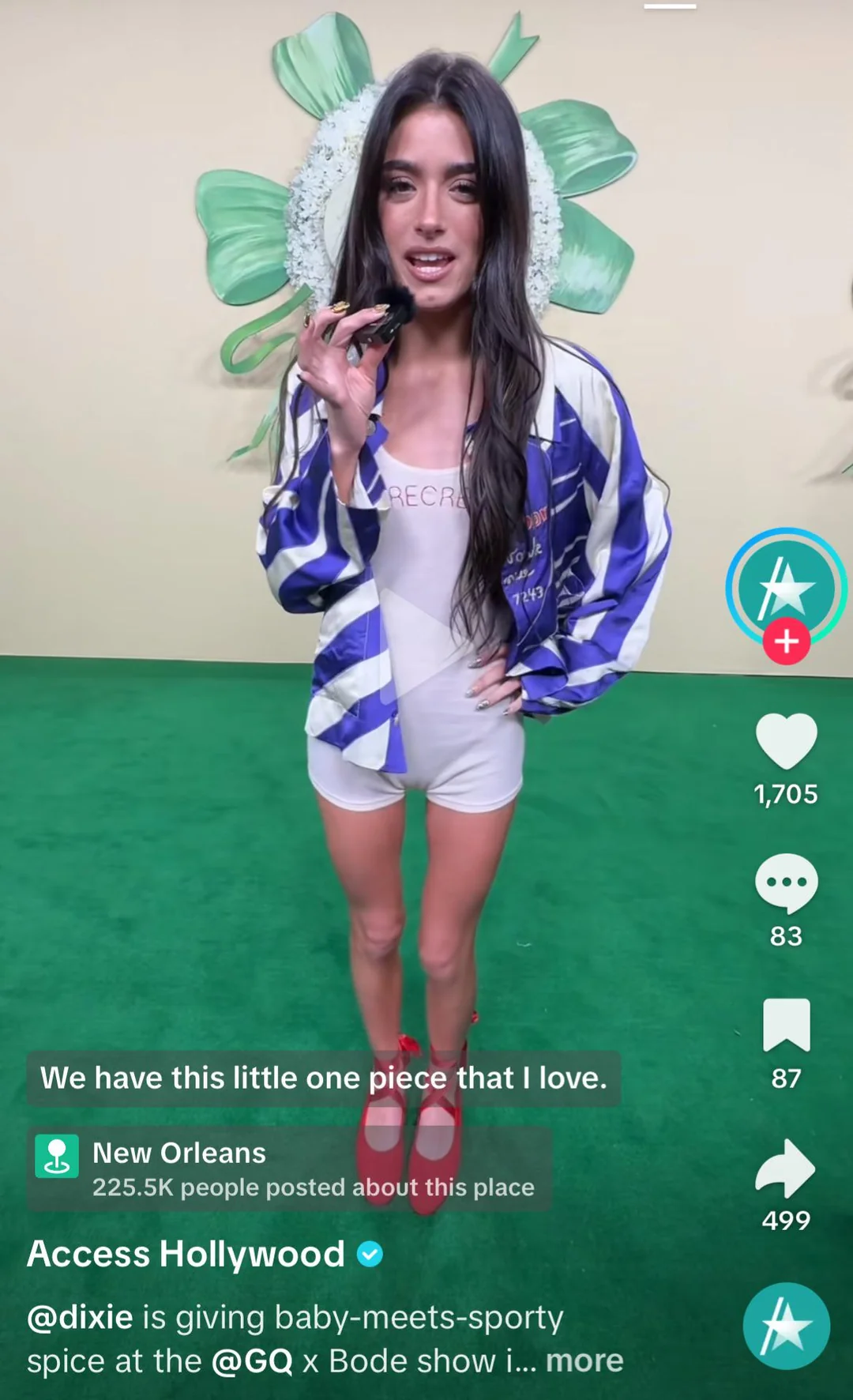 A young woman with long dark hair speaks into a microphone on a green carpet. She wears a blue and white jacket, white outfit, and red shoes. A large green floral backdrop is behind her. Various social media icons appear on screen.