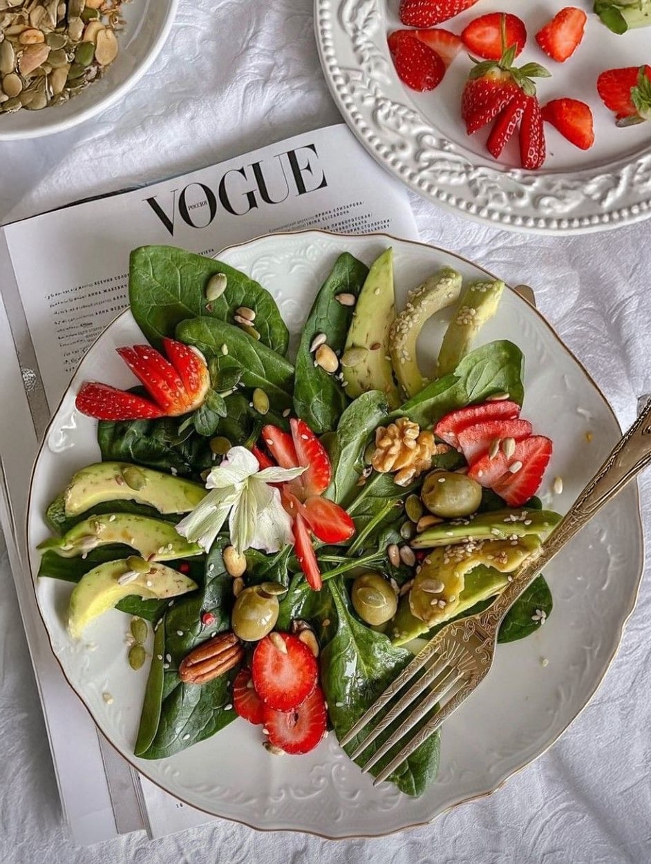 A fresh salad with spinach, avocado, strawberries, nuts, seeds, and olives is served on a white plate, sitting on a Vogue magazine, with another plate of strawberries and a bowl of seeds nearby.