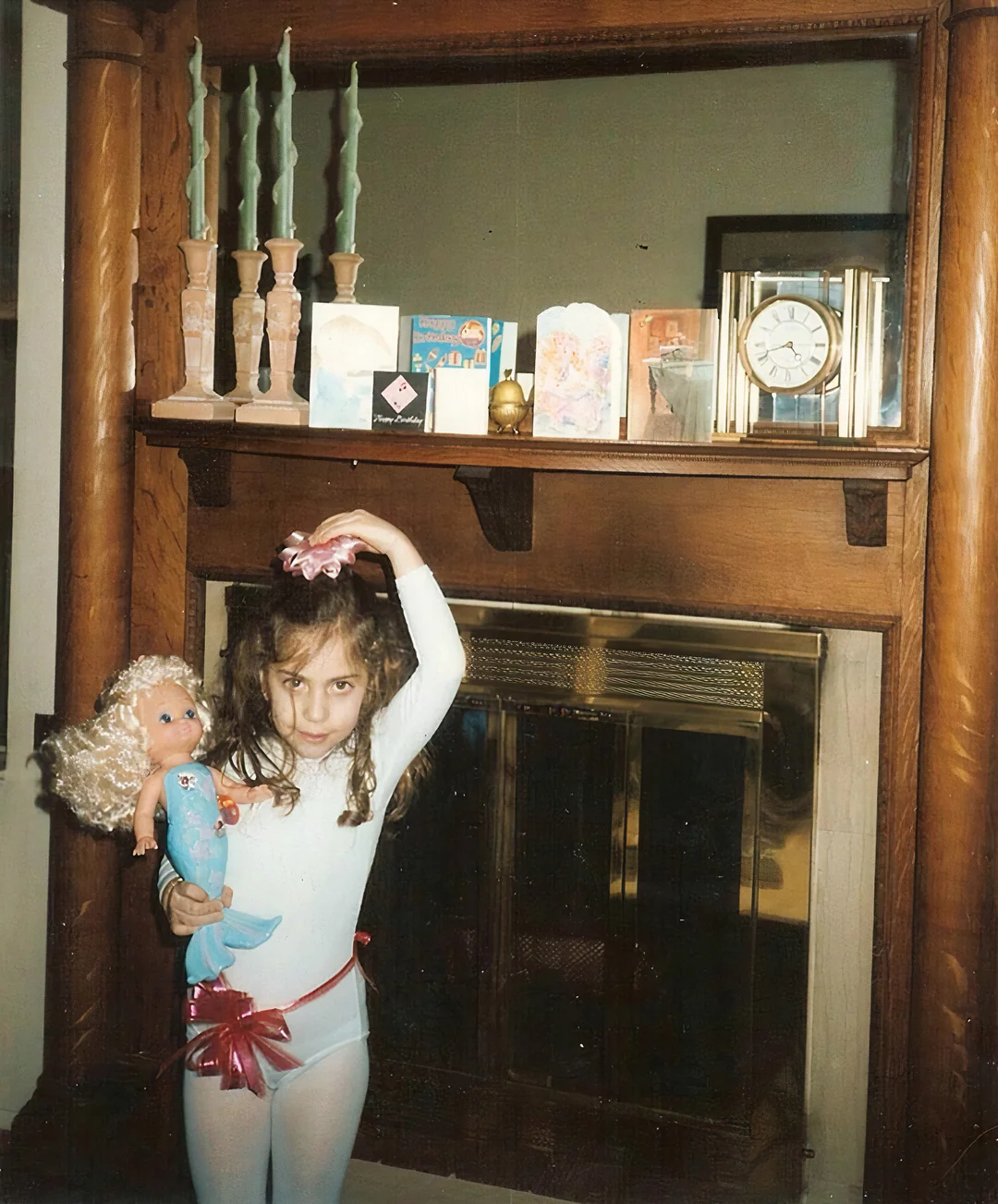 A young girl in a white leotard and pink tights holds a blonde mermaid doll and wears a pink scrunchie on her wrist. She poses in front of a wooden fireplace adorned with candles, greeting cards, and a clock.