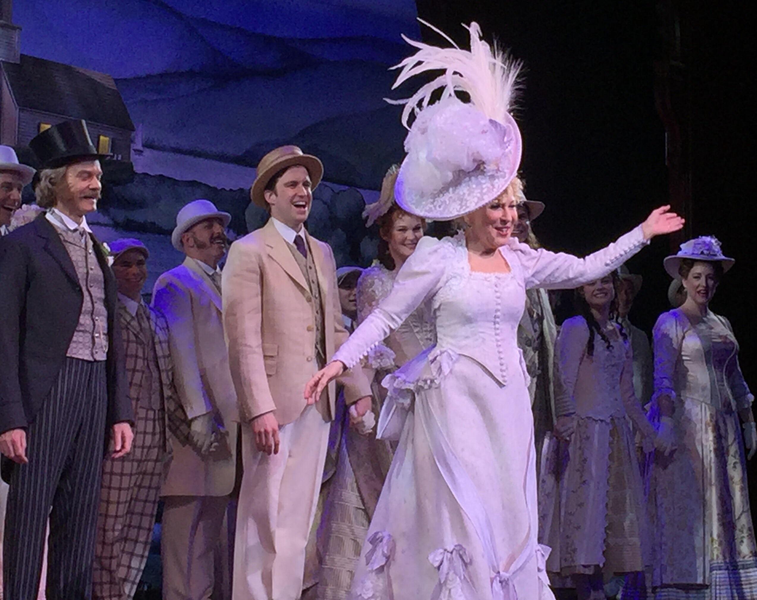 A woman in an elaborate white dress and large feathered hat stands center stage, smiling with arms outstretched, surrounded by people in vintage costumes, suggesting a theatrical performance.