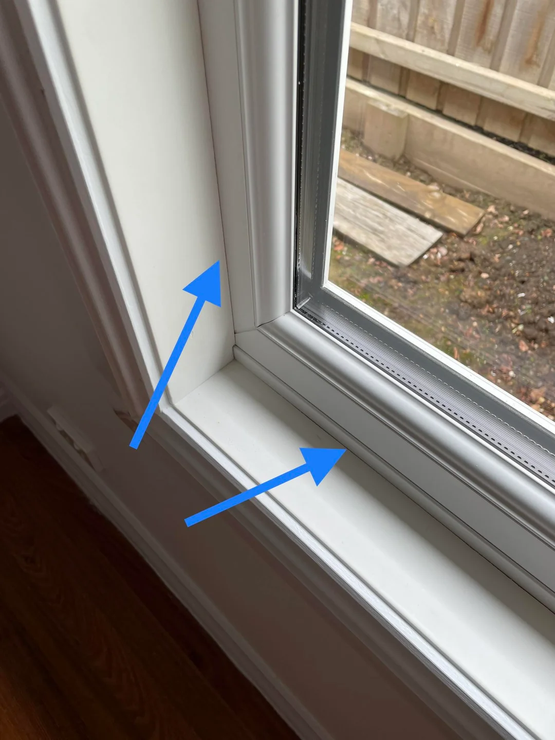 A close-up of a window frame with two blue arrows pointing to the bottom corner, highlighting the trim and sill detail. A wooden fence and yard are visible outside the window.