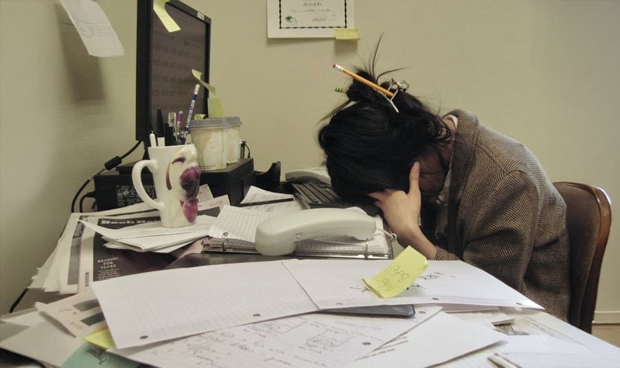 A person sits at a cluttered desk, head in hands, appearing stressed. Papers, a mug, a phone, and a pencil in their hair are visible. The computer monitor is on, and sticky notes are scattered around.