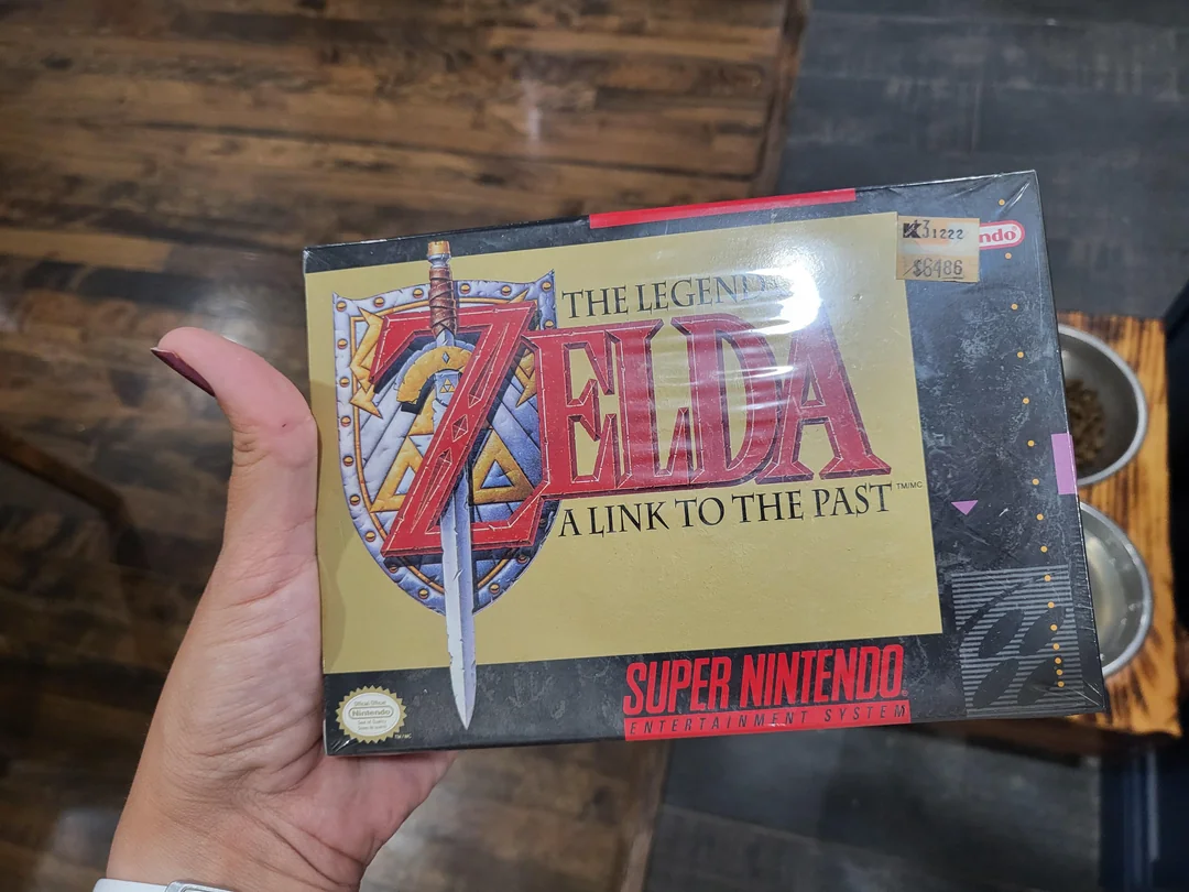 A hand holds a boxed copy of "The Legend of Zelda: A Link to the Past" for the Super Nintendo Entertainment System, featuring the game’s logo and sword on the cover.