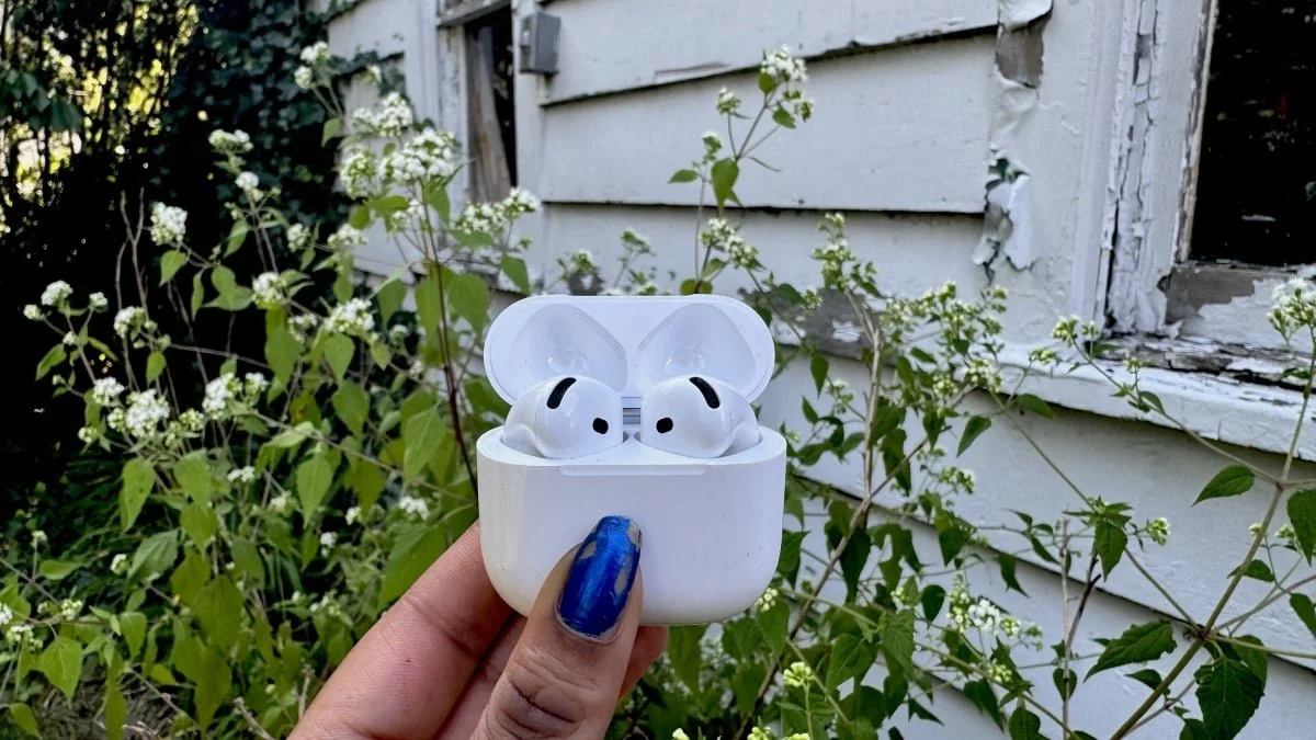 A hand with blue-painted nails holds a white case containing wireless earbuds outdoors, with green plants and a weathered white wooden building in the background.