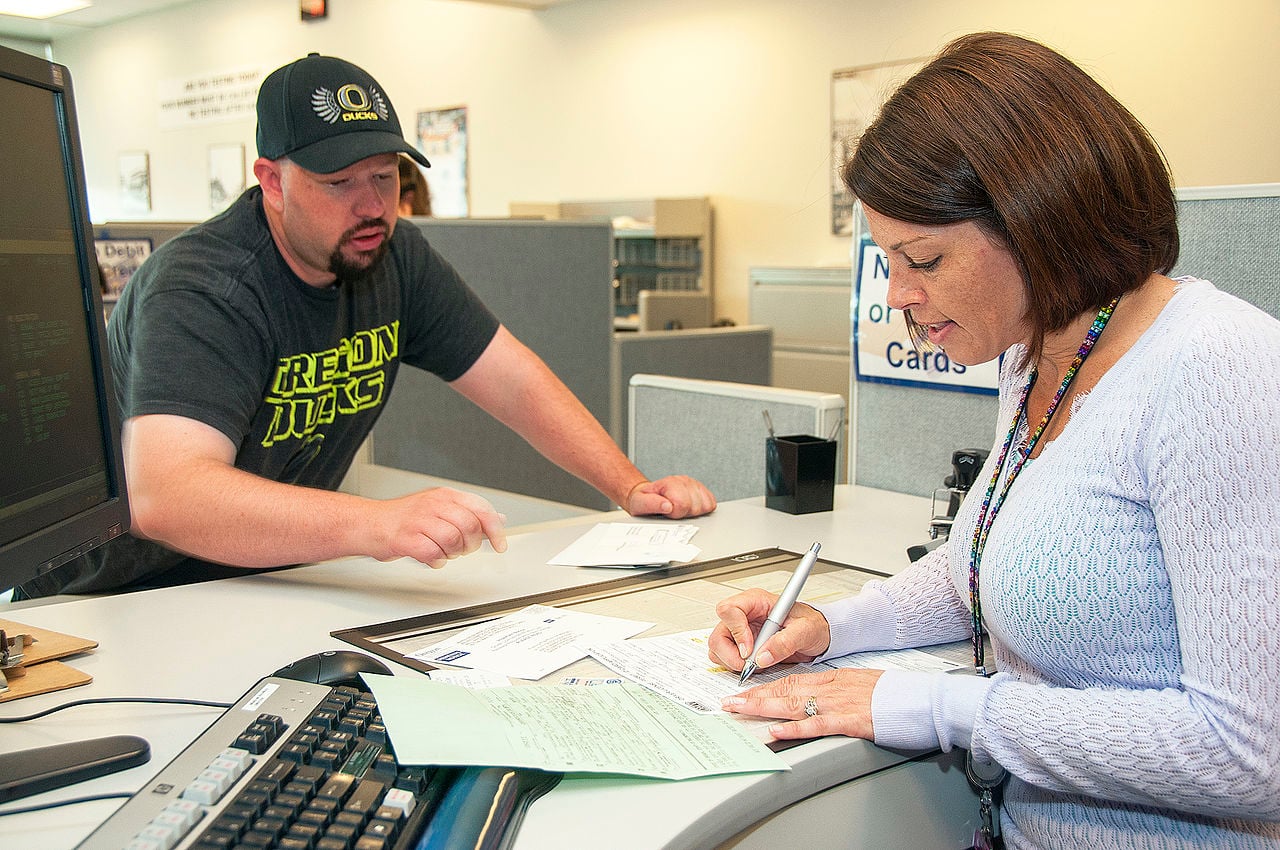 A woman fills out paperwork at a counter while a man in an Oregon Ducks shirt speaks to her. They are in an office setting with computers and documents on the desk.