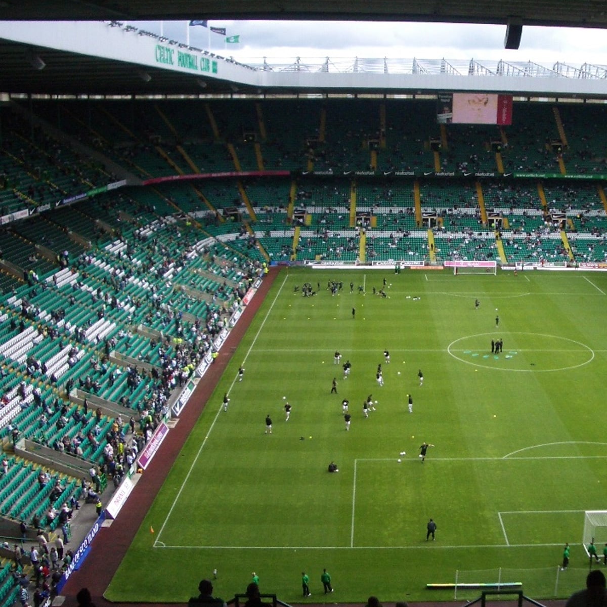 A large soccer stadium with green seats, partially filled with spectators, and players warming up on the field. The roof covers part of the stands, and the atmosphere is calm before a match.