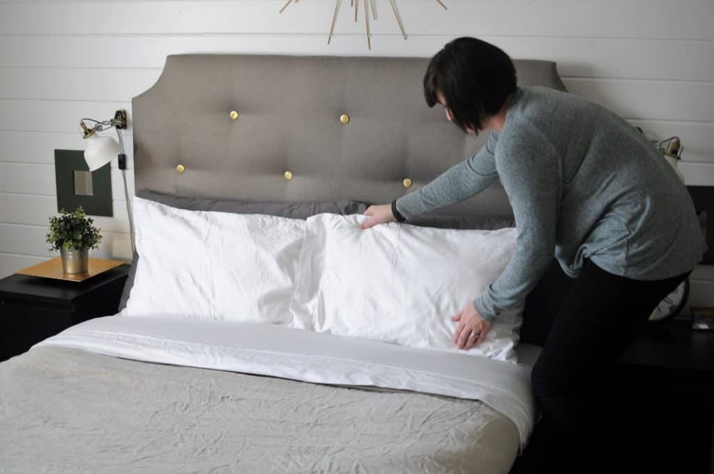 A person with short dark hair, wearing a gray long-sleeve shirt, arranges white pillows on a neatly made bed with a tufted gray headboard in a modern bedroom.